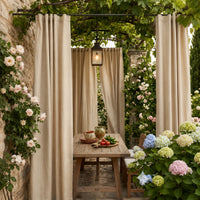 Beige Sunbrella outdoor curtains framing a vine-covered garden dining nook with stone wall, climbing roses, and hydrangeas — custom pergola curtains