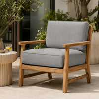 A modern outdoor armchair with light gray cushions and a natural wood frame, positioned on a patio next to a small round side table.