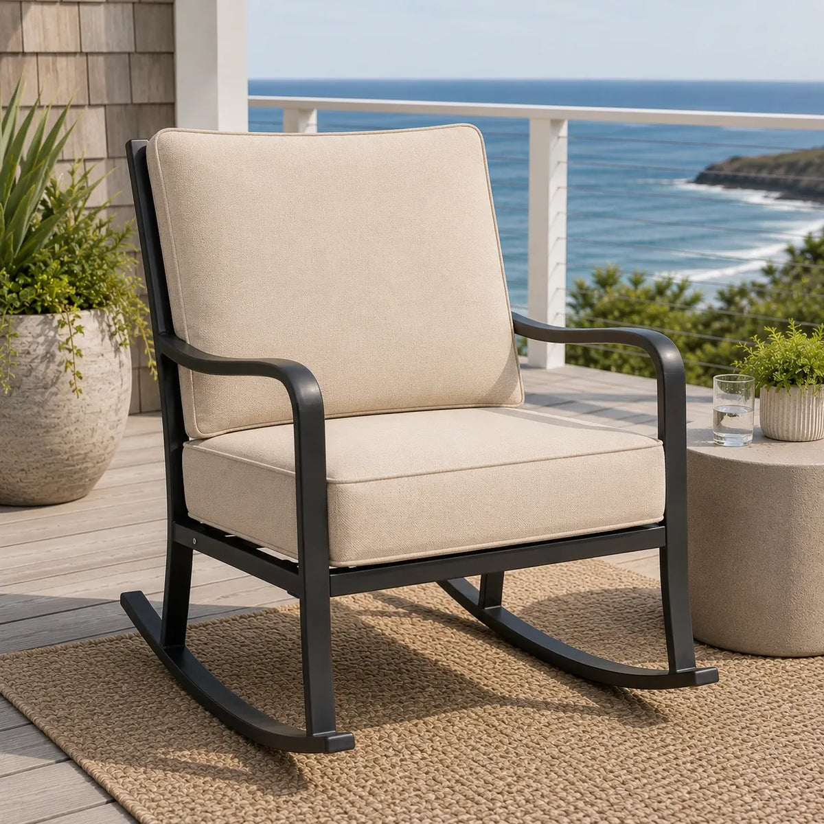 Outdoor rocking chair with beige cushions and a black metal frame, placed on a woven rug on a wooden deck with an ocean view background.