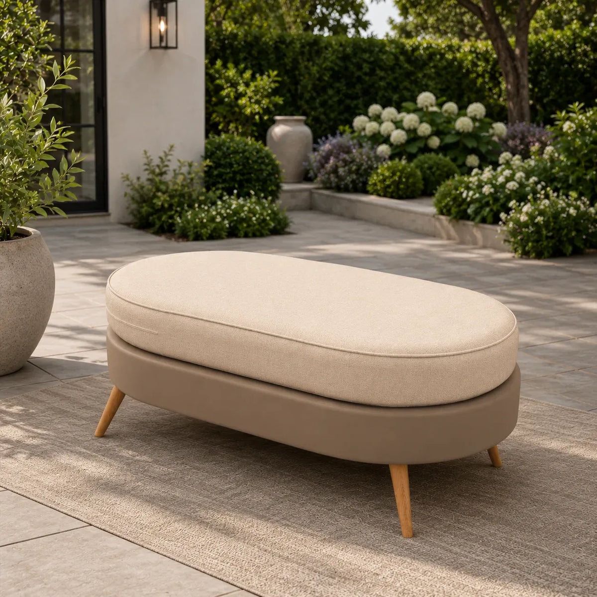 An outdoor oval ottoman with a light beige cushioned top and a taupe upholstered base on tapered wooden legs, placed on a patio in a landscaped garden setting.