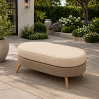 An outdoor oval ottoman with a light beige cushioned top and a taupe upholstered base on tapered wooden legs, placed on a patio in a landscaped garden setting.