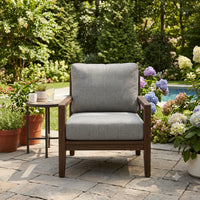 A single outdoor patio armchair with a dark brown wooden frame and light gray textured cushions, set on a stone patio in a lush garden with a side table next to it.