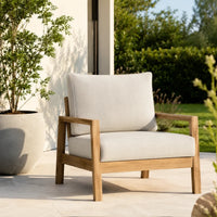 A modern outdoor armchair with light beige cushions and a natural wood frame, positioned on a light stone patio with a potted plant and green garden in the background.