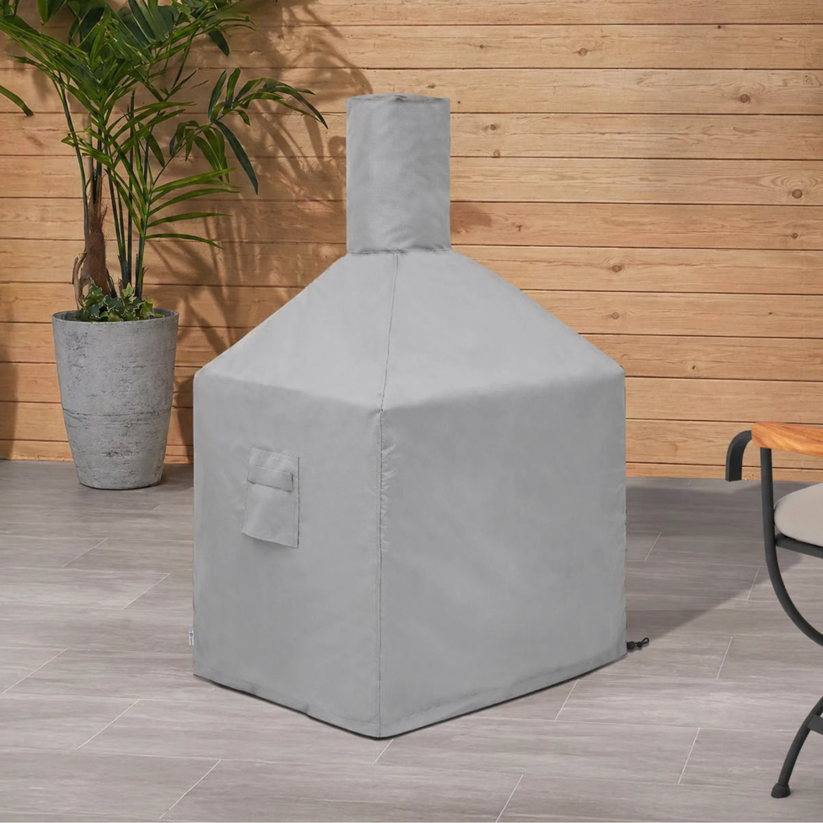 Custom Square Fire Pit Chiminea Cover in light gray, shown on a patio with wood panel wall and tropical plant