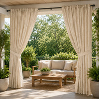 A pair of textured cream-beige patio curtains hanging on a black rod over an open porch, framing a furnished outdoor seating area with a wooden coffee table, sofa, and potted plants.