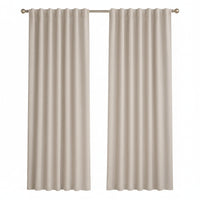 A pair of light beige curtain panels hanging on a silver rod in a bright, modern room with a window, a small wooden side table with a vase, and a light-colored armchair.