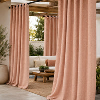 A set of dusty rose pink textured outdoor curtains hanging on a black rod in a patio setting with a white sofa, wooden table, and plants.