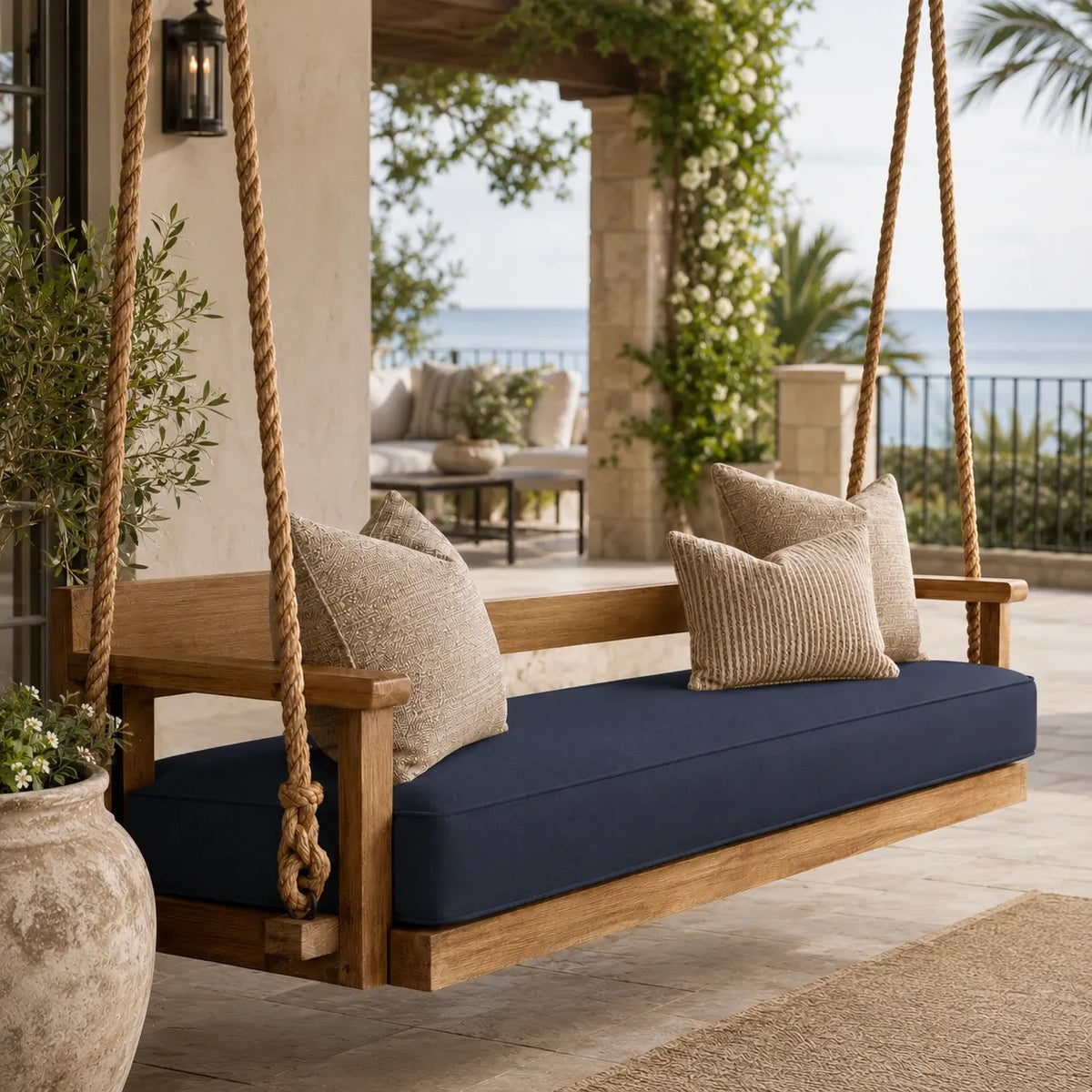 A wooden hanging porch swing with a navy blue cushion and beige textured pillows, set on a stone patio with an ocean view in the background.