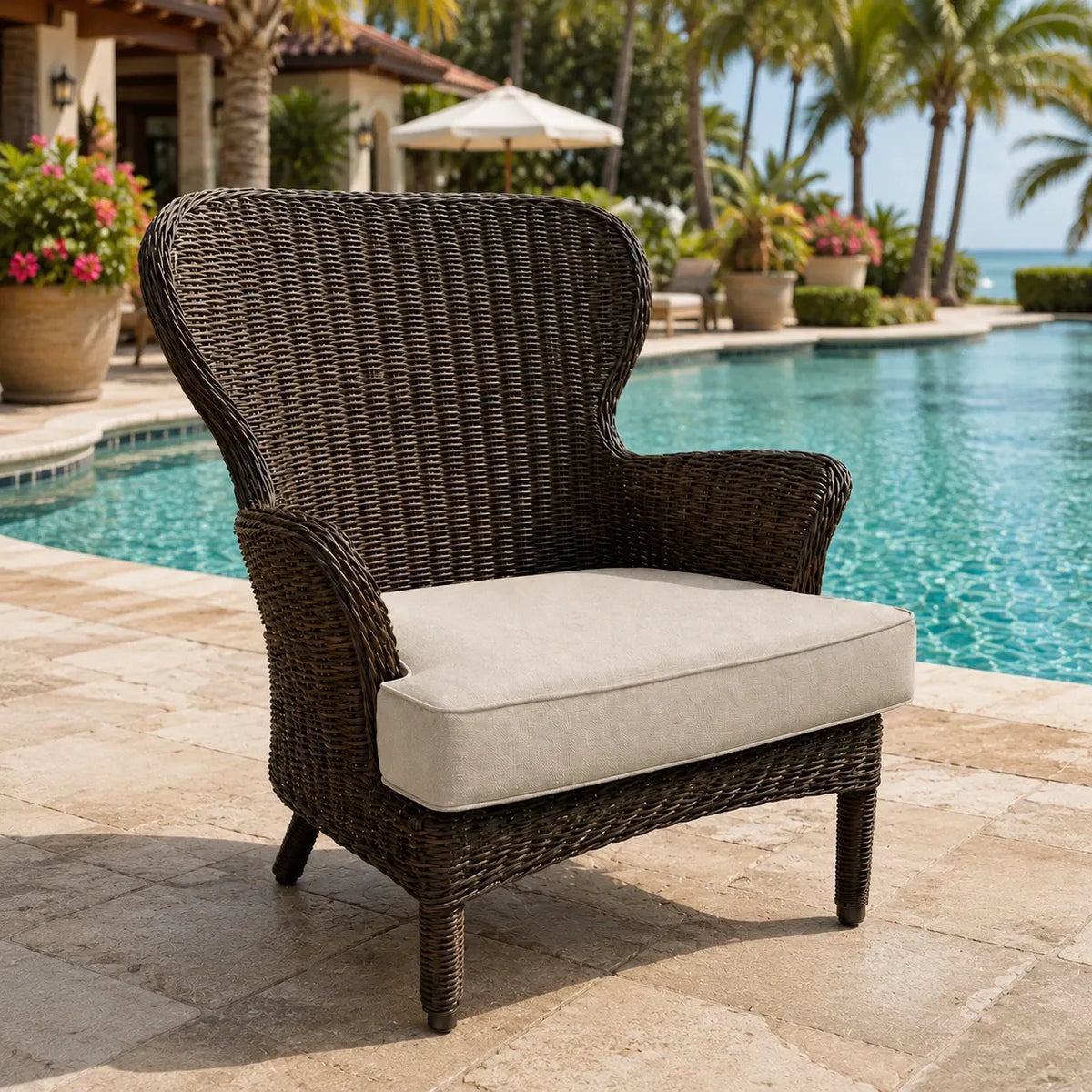 A dark brown woven outdoor armchair with a light beige cushion, positioned on a stone patio next to a swimming pool with a tropical resort background.
