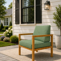 A wooden outdoor patio armchair with sage green cushions, positioned on a light beige woven rug in front of a white house with black shutters.