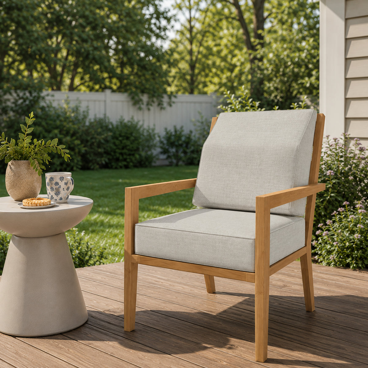 A modern outdoor armchair with a light beige linen cushion and a natural wood frame, positioned on a wooden deck in a backyard setting next to a small side table.