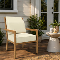 A wooden outdoor armchair with cream-colored cushions, set on a wooden deck next to a small side table with plants and a mug, in front of a house with white-framed glass doors.